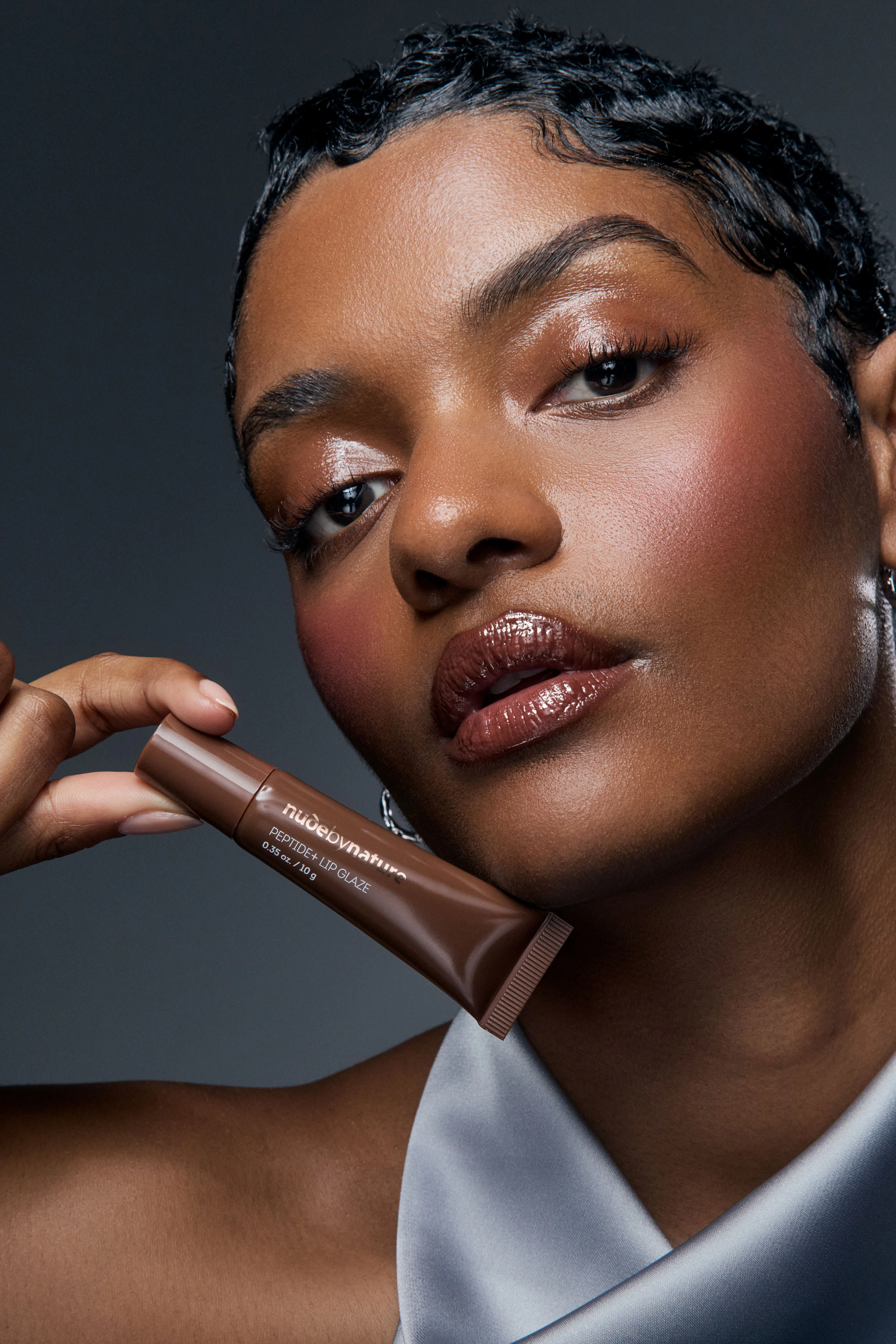 Woman wearing brown glossy lip glaze holding brown product tube near her face against a dark studio backdrop for an editorial style Nude by Nature beauty photography campaign
