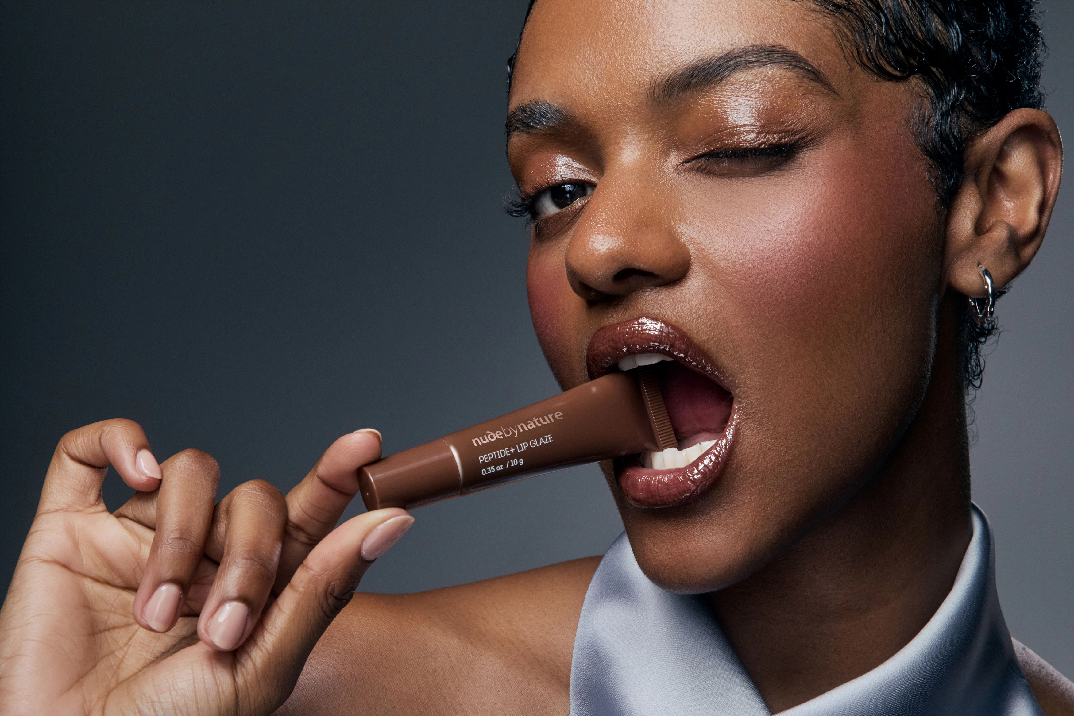 Woman wearing glossy brown lip balm holding a brown product tube between her teeth against a dark studio backdrop for a Nude by Nature beauty photography campaign