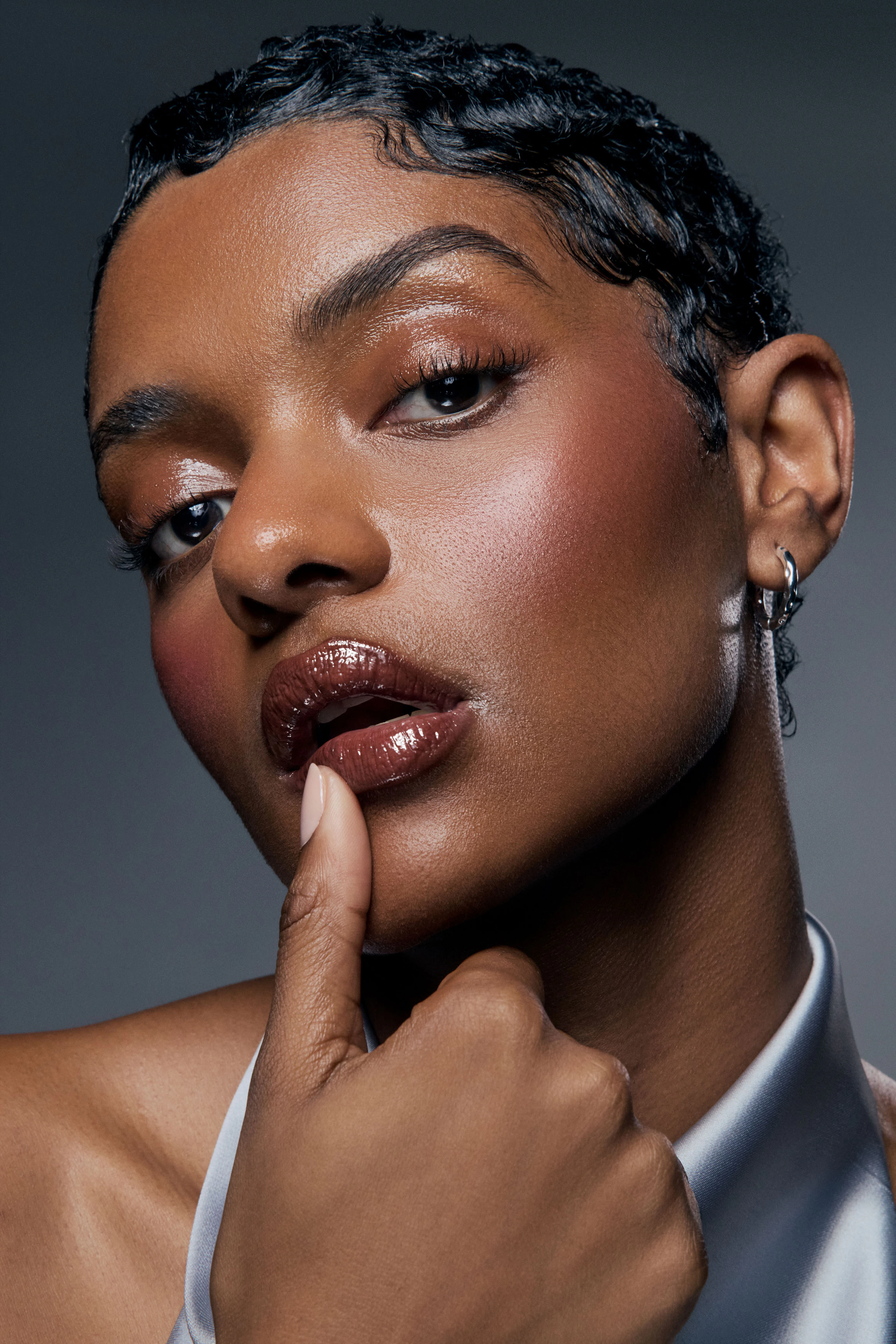 Woman with wet look hair wearing brown glossy lip glaze with thumb on lip against dark studio backdrop for an editorial style Nude by Nature beauty photography campaign
