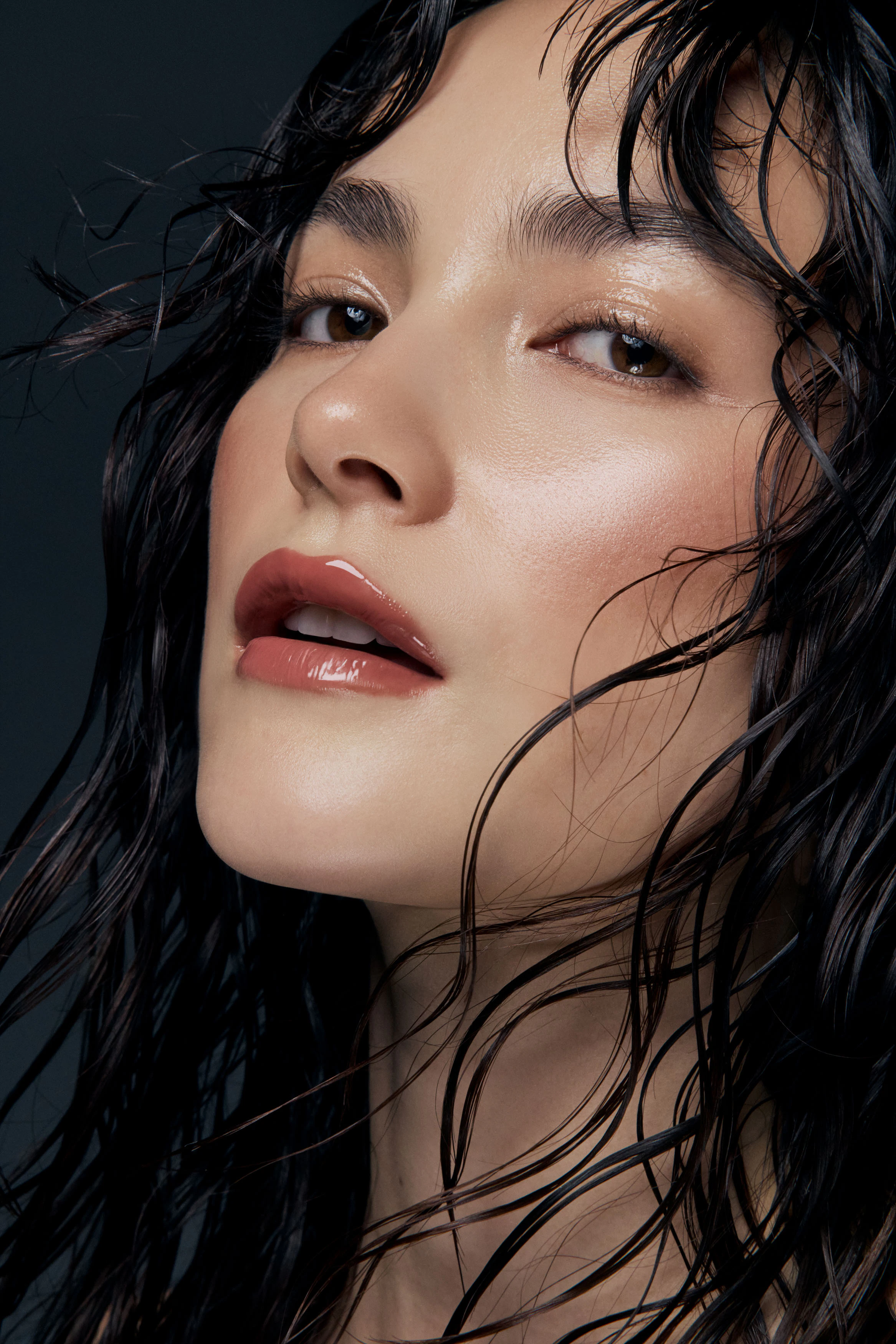 Close up shot of woman with wet look hair wearing pink glossy lip glaze against dark studio backdrop for an editorial style Nude by Nature beauty photography campaign
