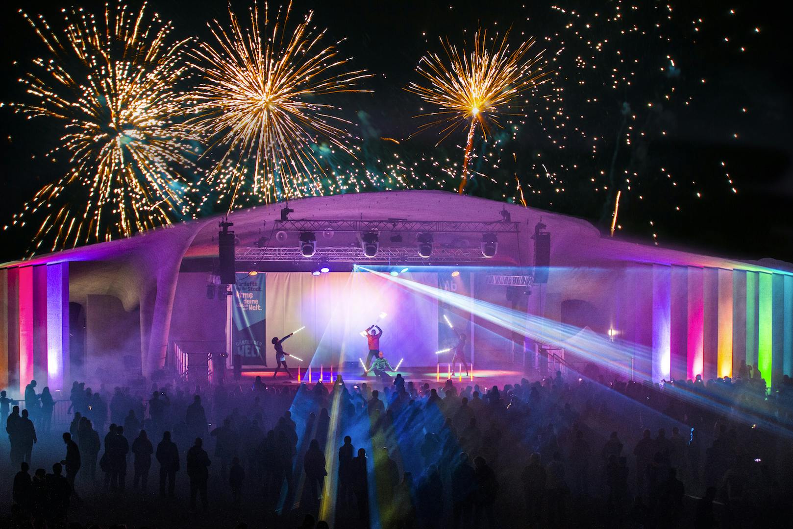 Outdoorbühne mit Artisten auf der Bühne und Feuerwerk und Lasershow im Hintergrund und vielen Zuschauern. 