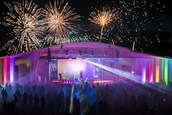 Outdoorbühne mit Artisten auf der Bühne und Feuerwerk und Lasershow im Hintergrund und vielen Zuschauern. 