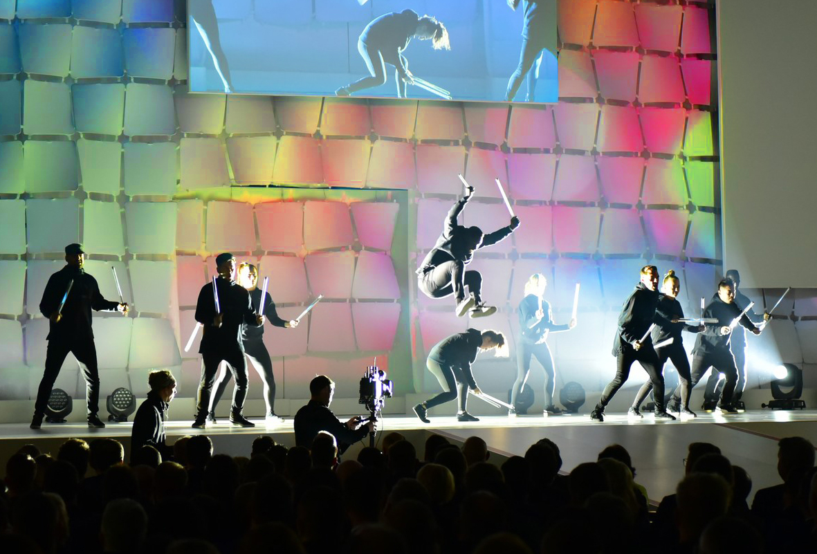 Lichtshow mit Breakdancern, die in einer Tanzformation stehen, ihre Arme heben und zwei LED-Leuchtstäbe voreinander kreuzen.