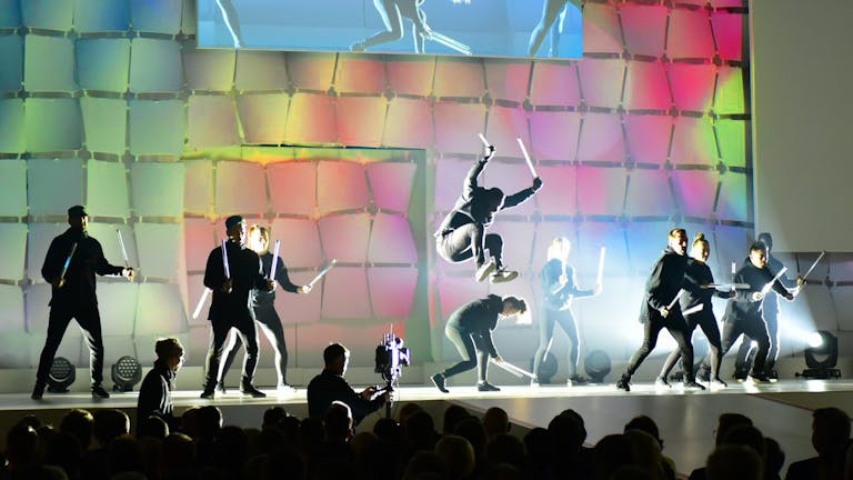 Lichtshow mit Breakdancern, die in einer Tanzformation stehen, ihre Arme heben und zwei LED-Leuchtstäbe voreinander kreuzen.