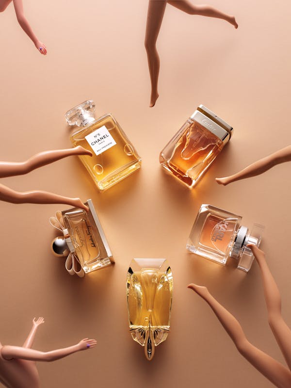 A group of five perfumes lie on a beige background. In front of them, the legs and hands of Barbie dolls dance.