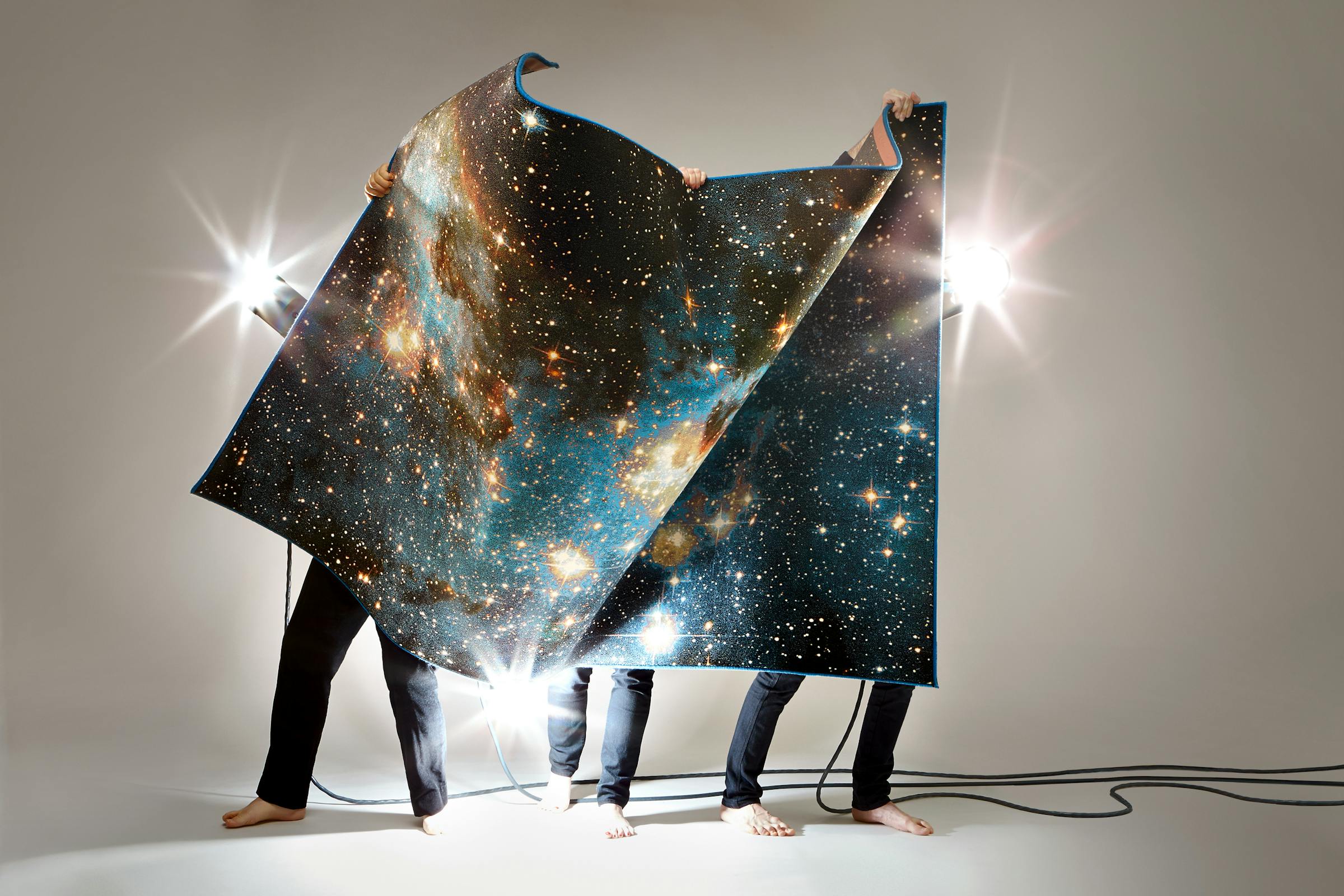 Three men in black trousers hold up a carpet with a print of the universe.