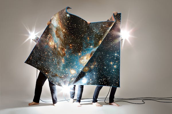 Three men in black trousers hold up a carpet with a print of the universe.