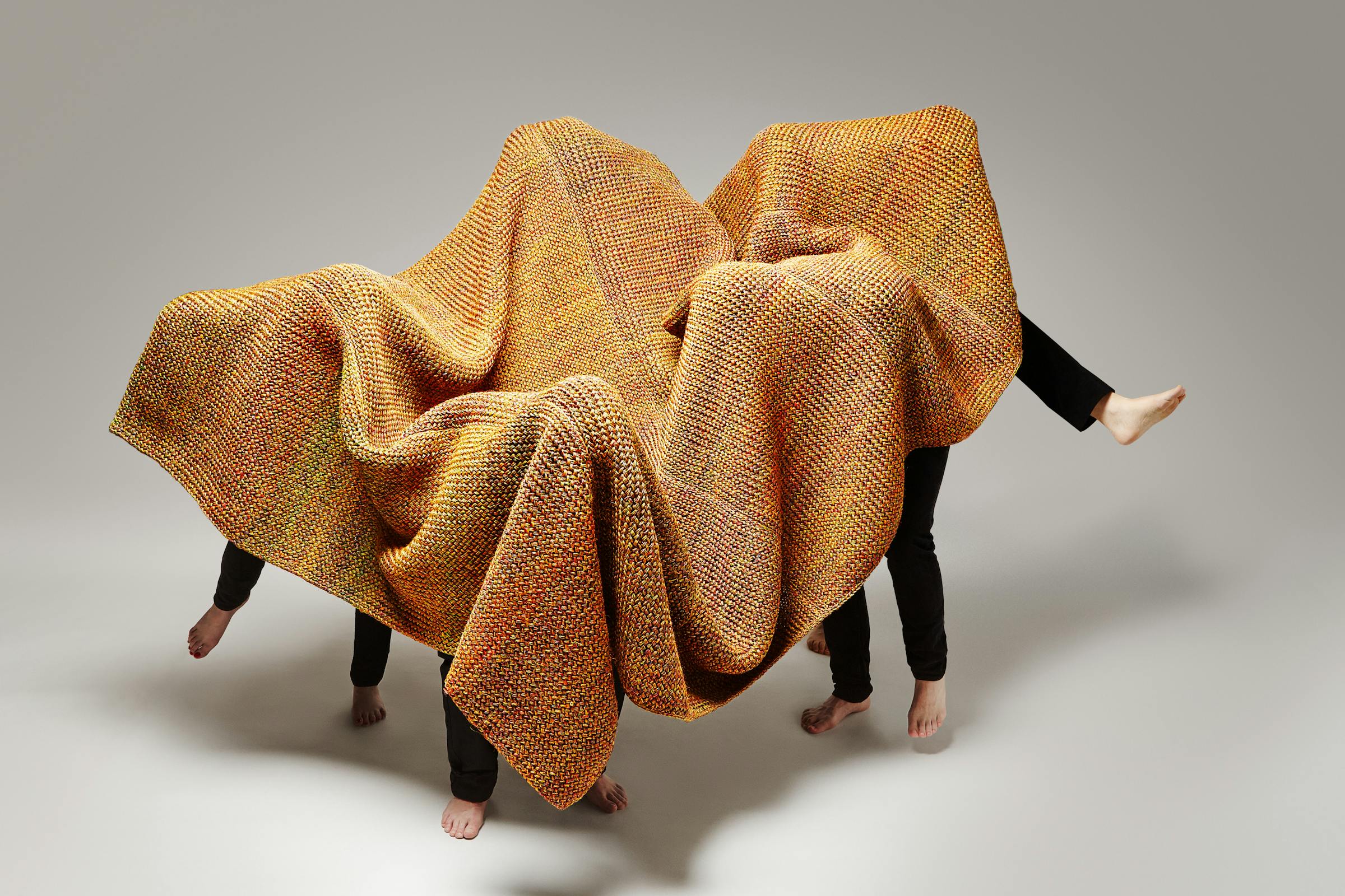 Four men in black trousers dance under a large knitted carpet in orange.