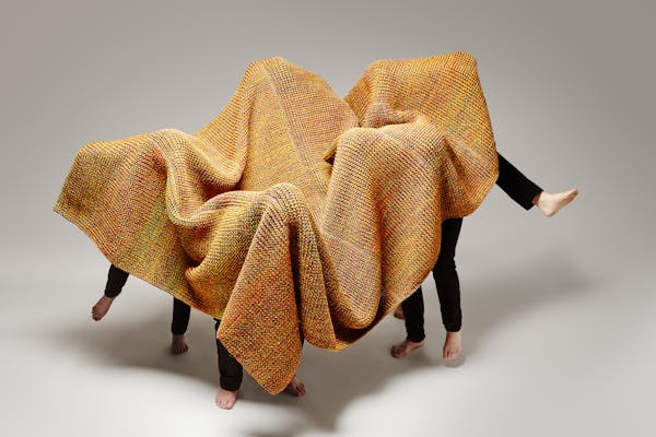 Four men in black trousers dance under a large knitted carpet in orange.