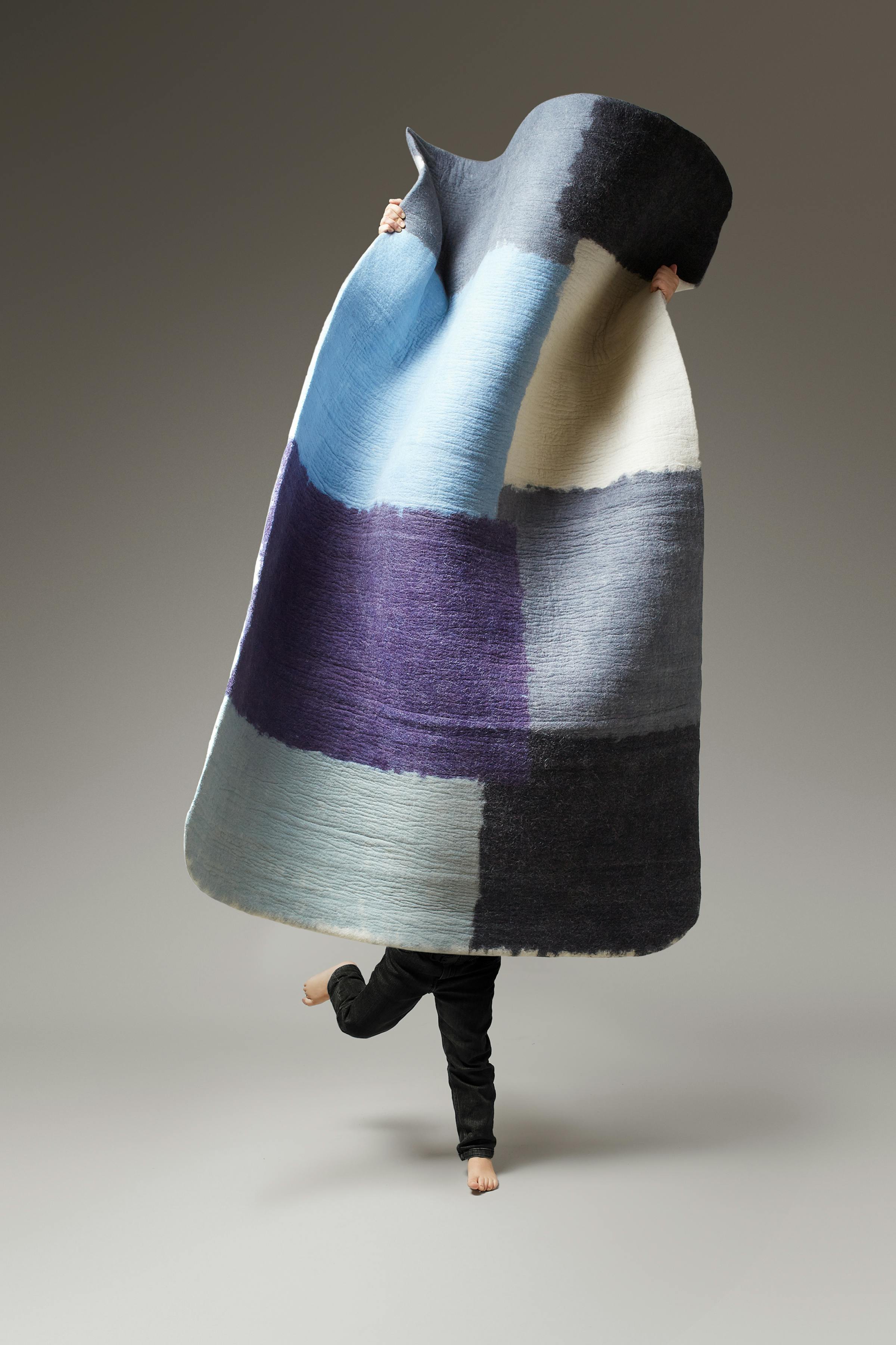 A little boy in black trousers is holding a large geometric rug.