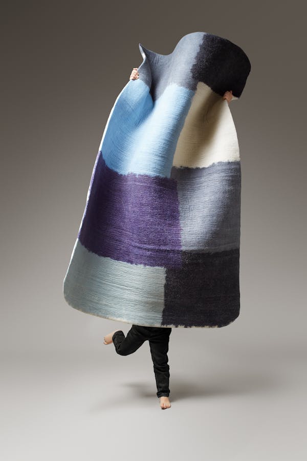 A little boy in black trousers is holding a large geometric rug.