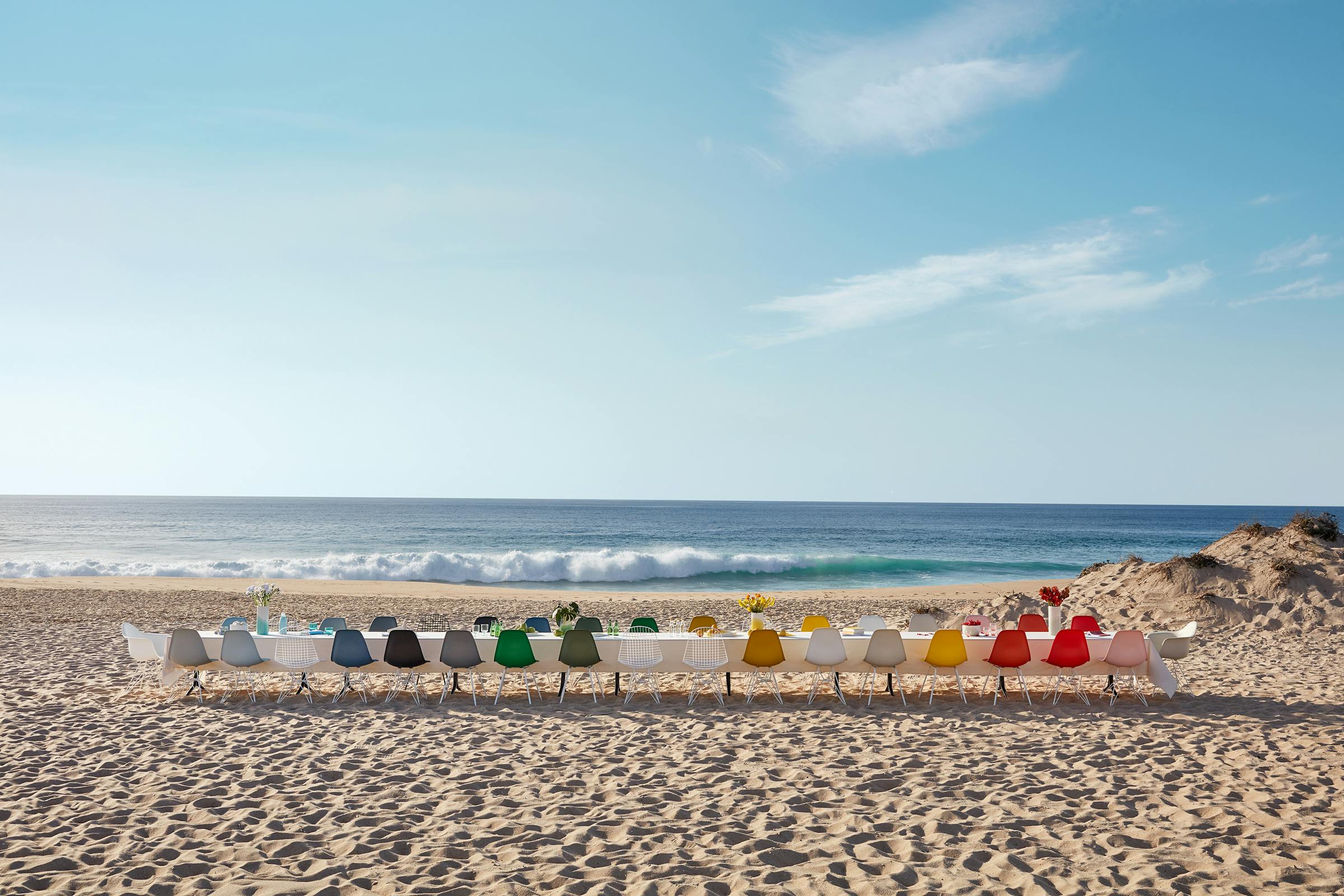 Long dining table with white tablecloth and Eames shell chairs in colours on a sunny day at the beach