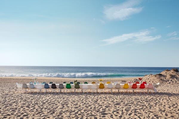 Long dining table with white tablecloth and Eames shell chairs in colours on a sunny day at the beach