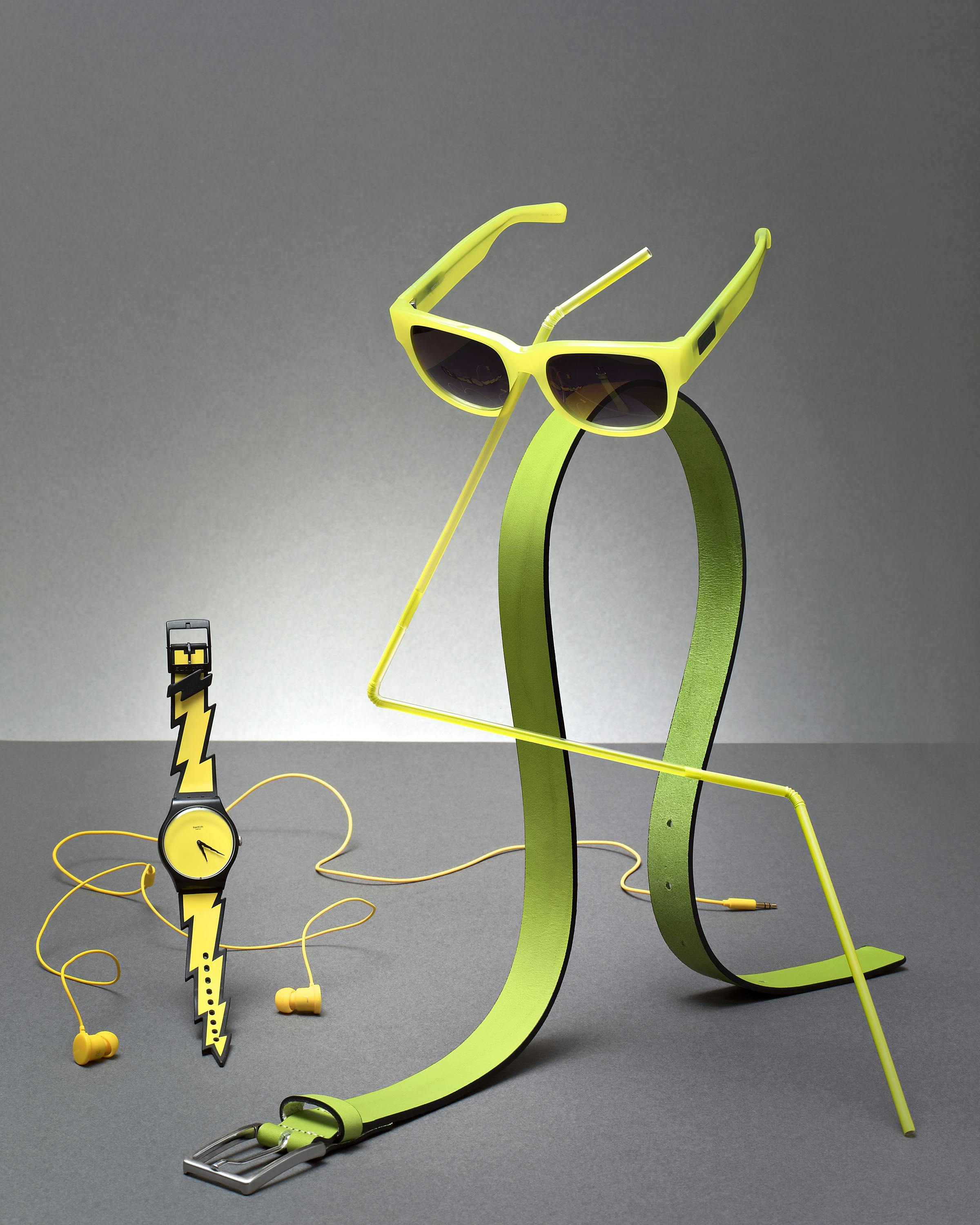 Sunglasses, a belt and a lightning bolt-shaped watch in green and yellow, sculpturally arranged together.