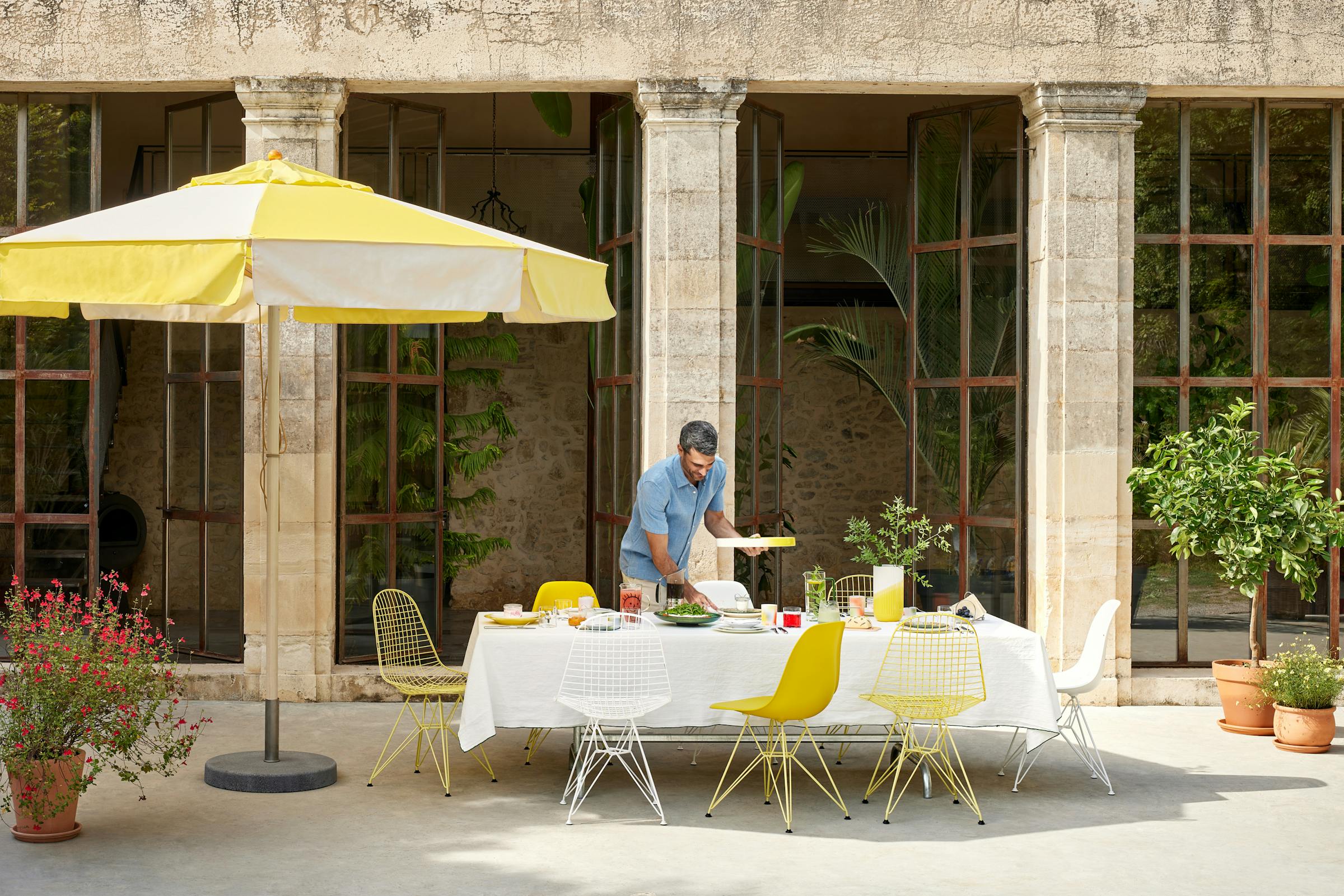 Lunch table in summer with a large yellow parasol and yellow Eames chairs. A man in a light blue denim shirt is bringing food
