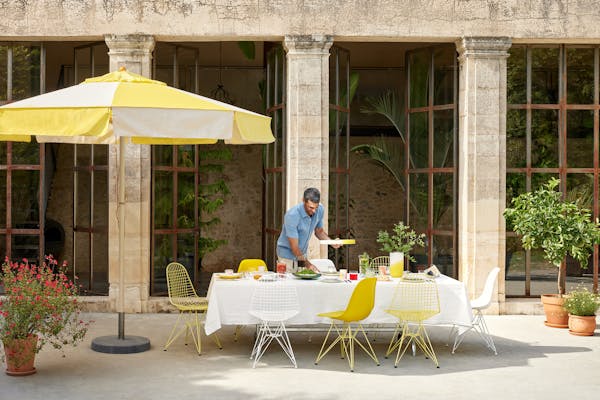 Lunch table in summer with a large yellow parasol and yellow Eames chairs. A man in a light blue denim shirt is bringing food