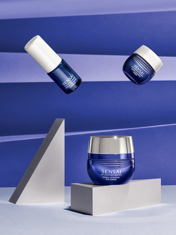 Three dark blue beauty products on silver cubes in front of a violet paper background