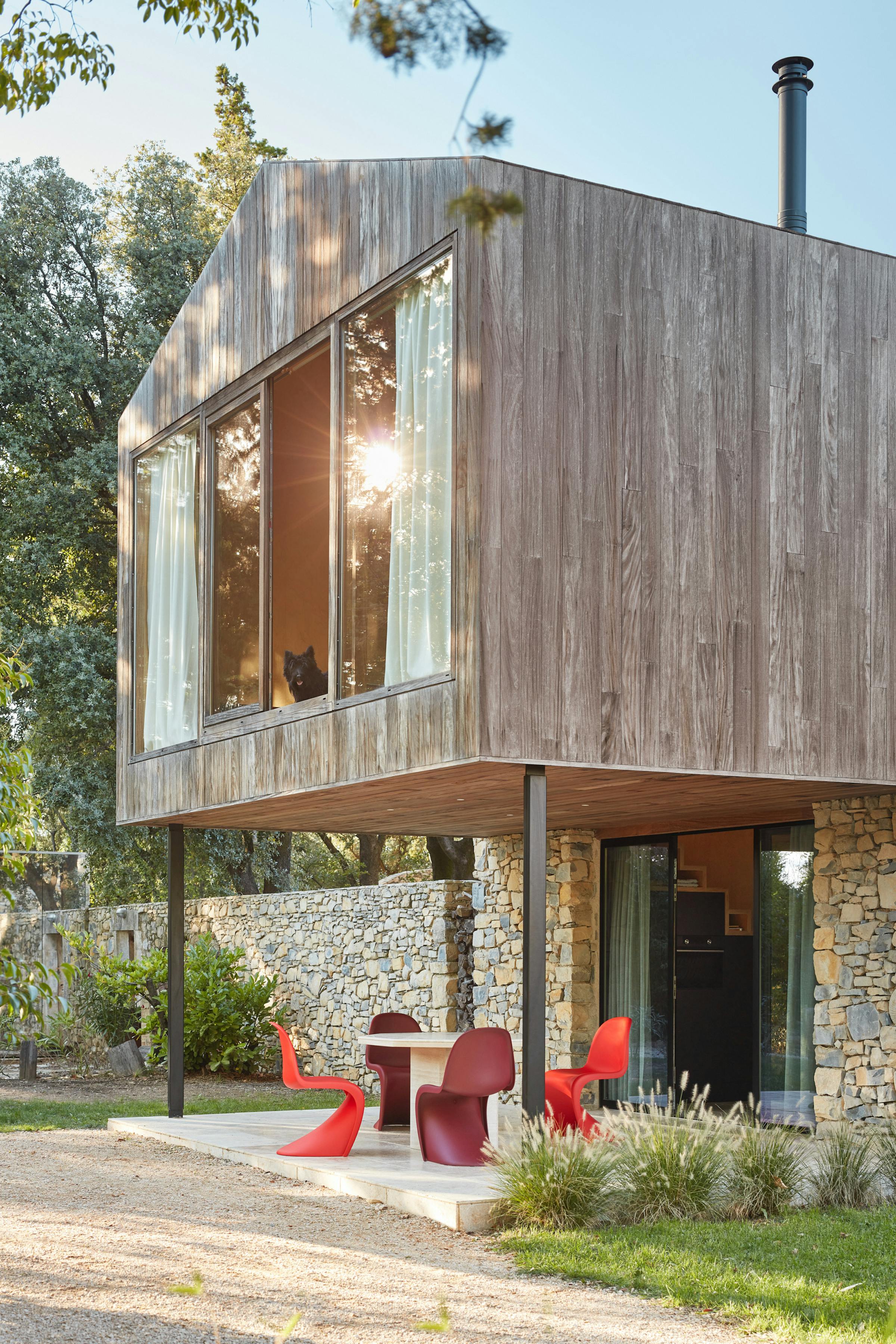 four red Panton chairs on the terrace of a modern wooden house with a dog looking out of the window