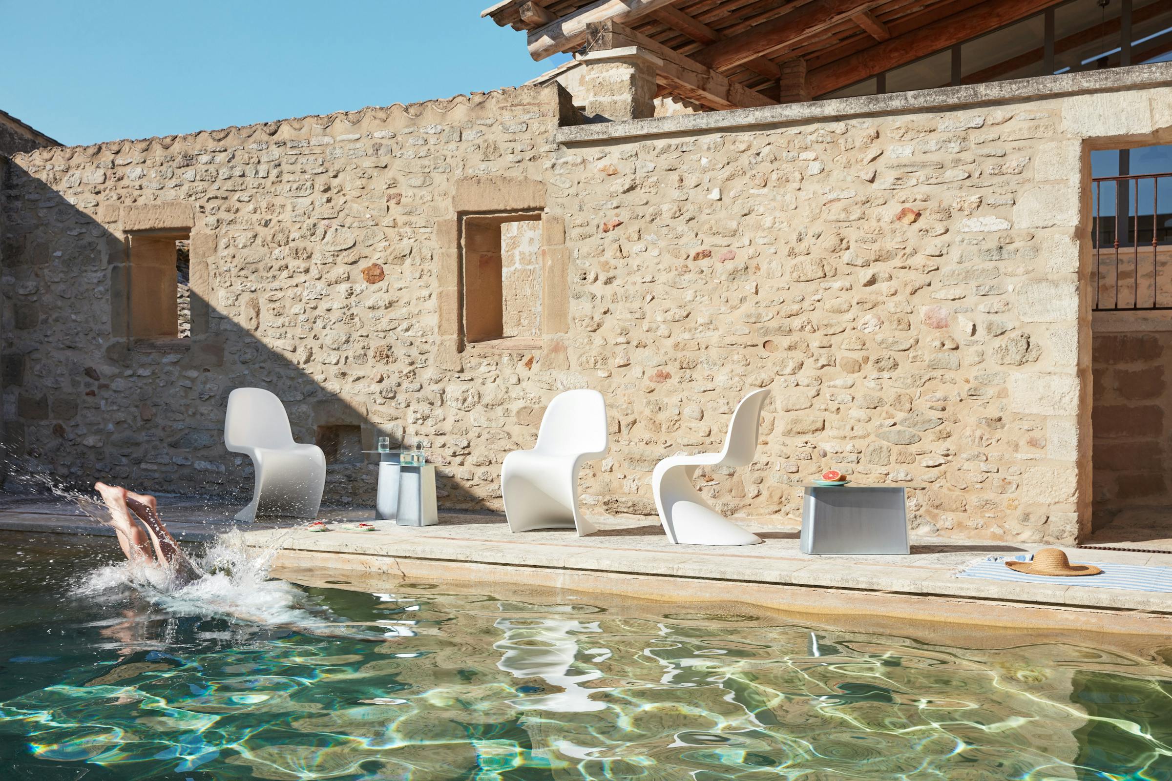 Three white Panton chairs stand in front of a stone wall by a pool in bright sunshine. A man jumps into the pool.