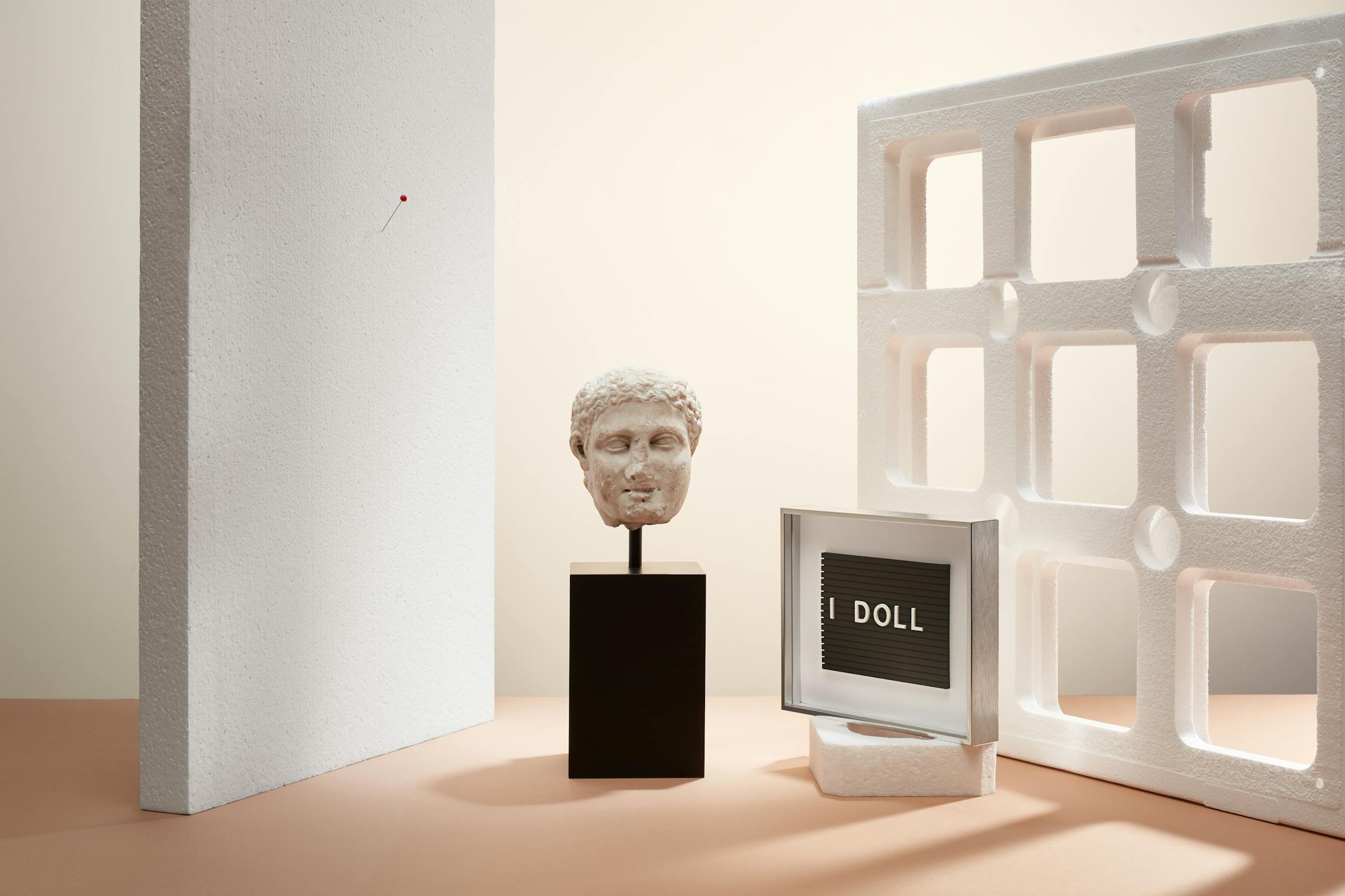 Ancient greek head and the text lines "I Doll" in a set design made out of white styrofoam objects