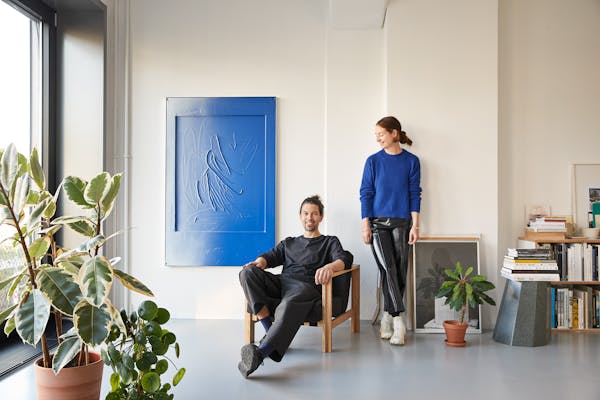 Young couple in front of a white wall with a bright blue picture. The woman is wearing a jumper in the same colour.