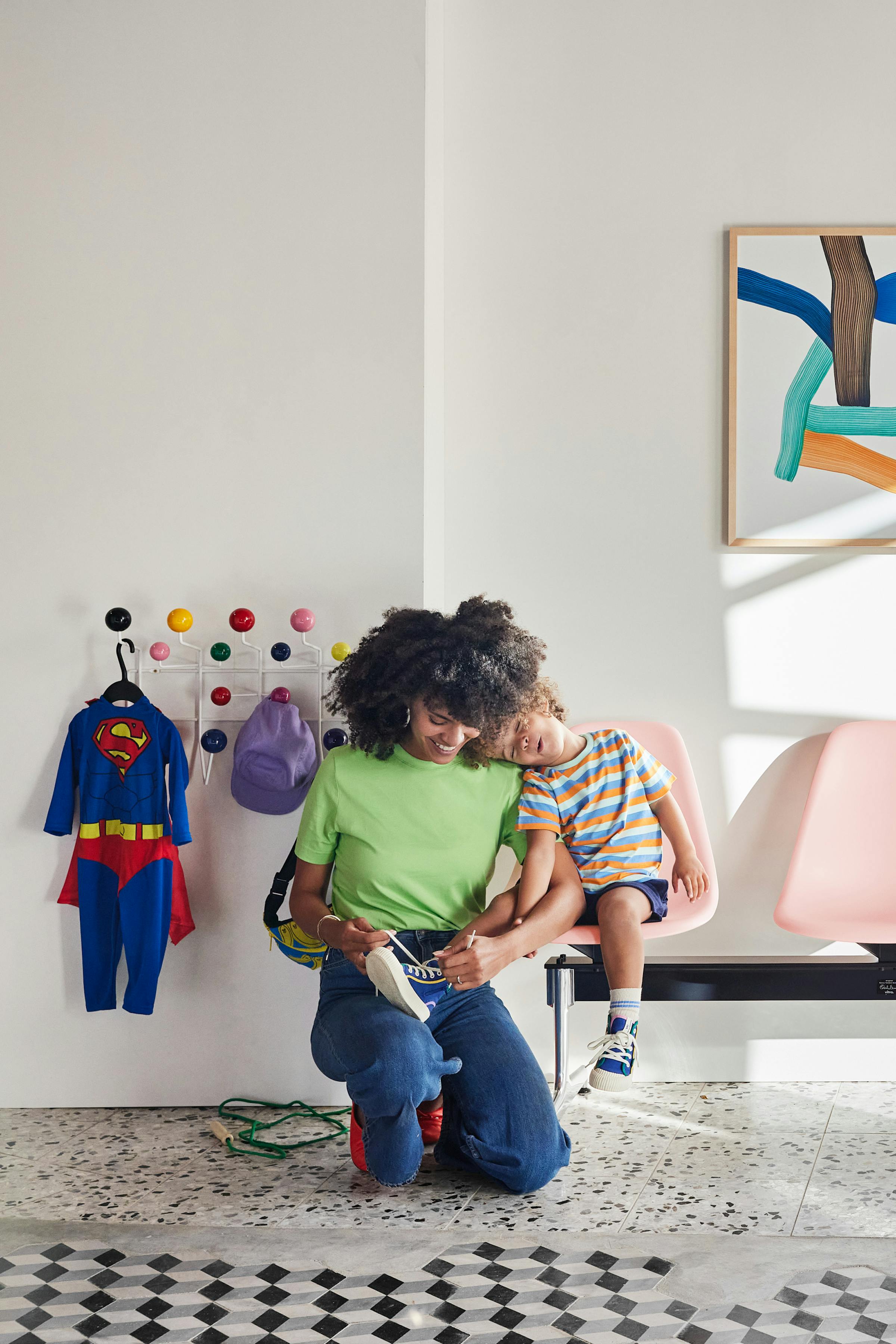 A young mother puts on her son's shoes as he sits on an Eames bench. On the wall  the Eames Hang it All is mounted, on it hangs a Superman costume