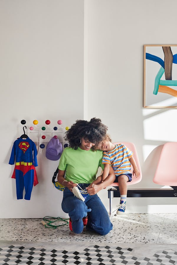 A young mother puts on her son's shoes as he sits on an Eames bench. On the wall  the Eames Hang it All is mounted, on it hangs a Superman costume
