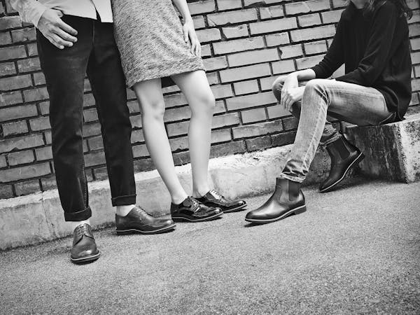 Three young people wearing black leather shoes in front of a brick wall