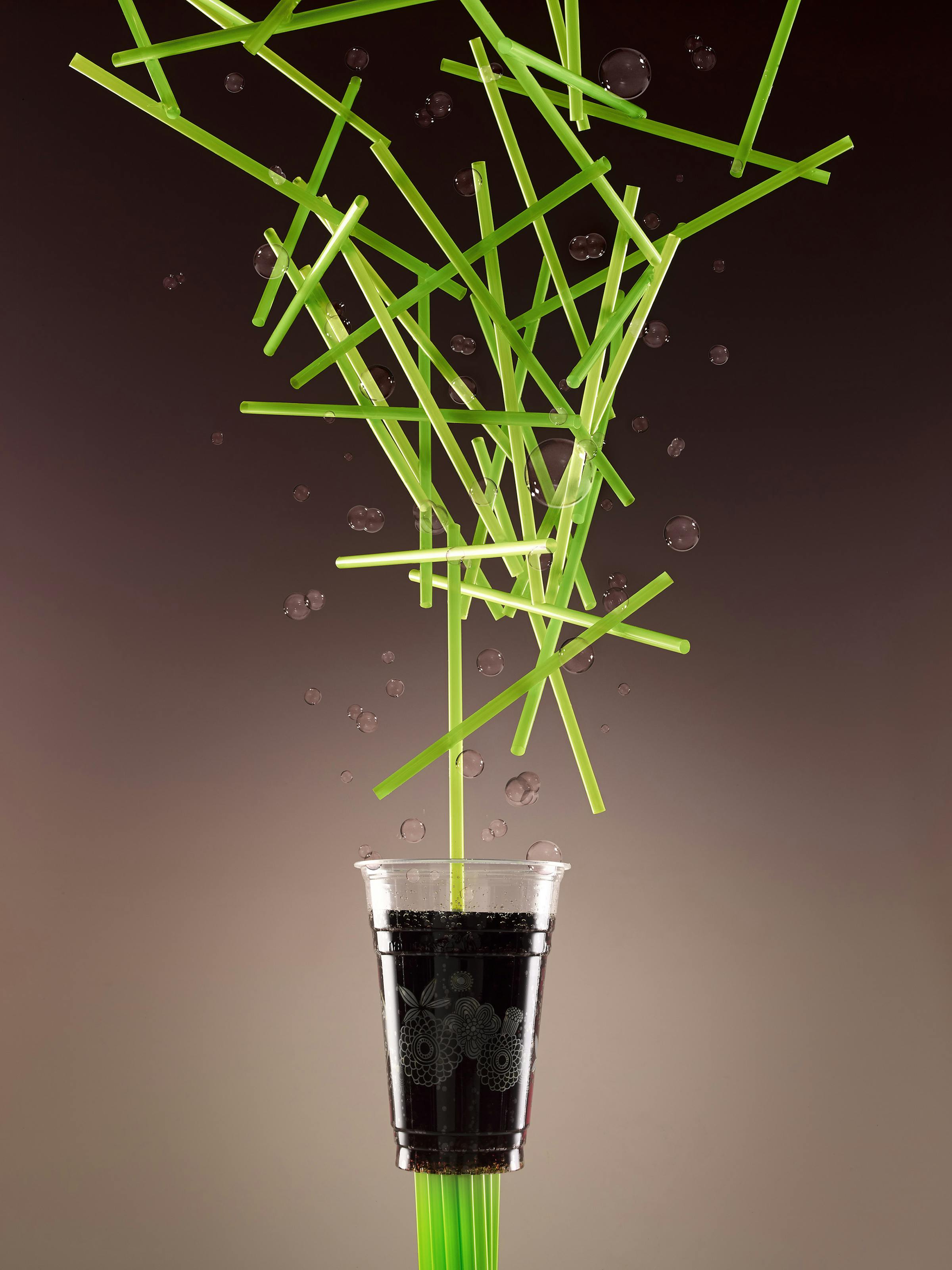 Plastic cup in front of a brown background with many green straws falling from above