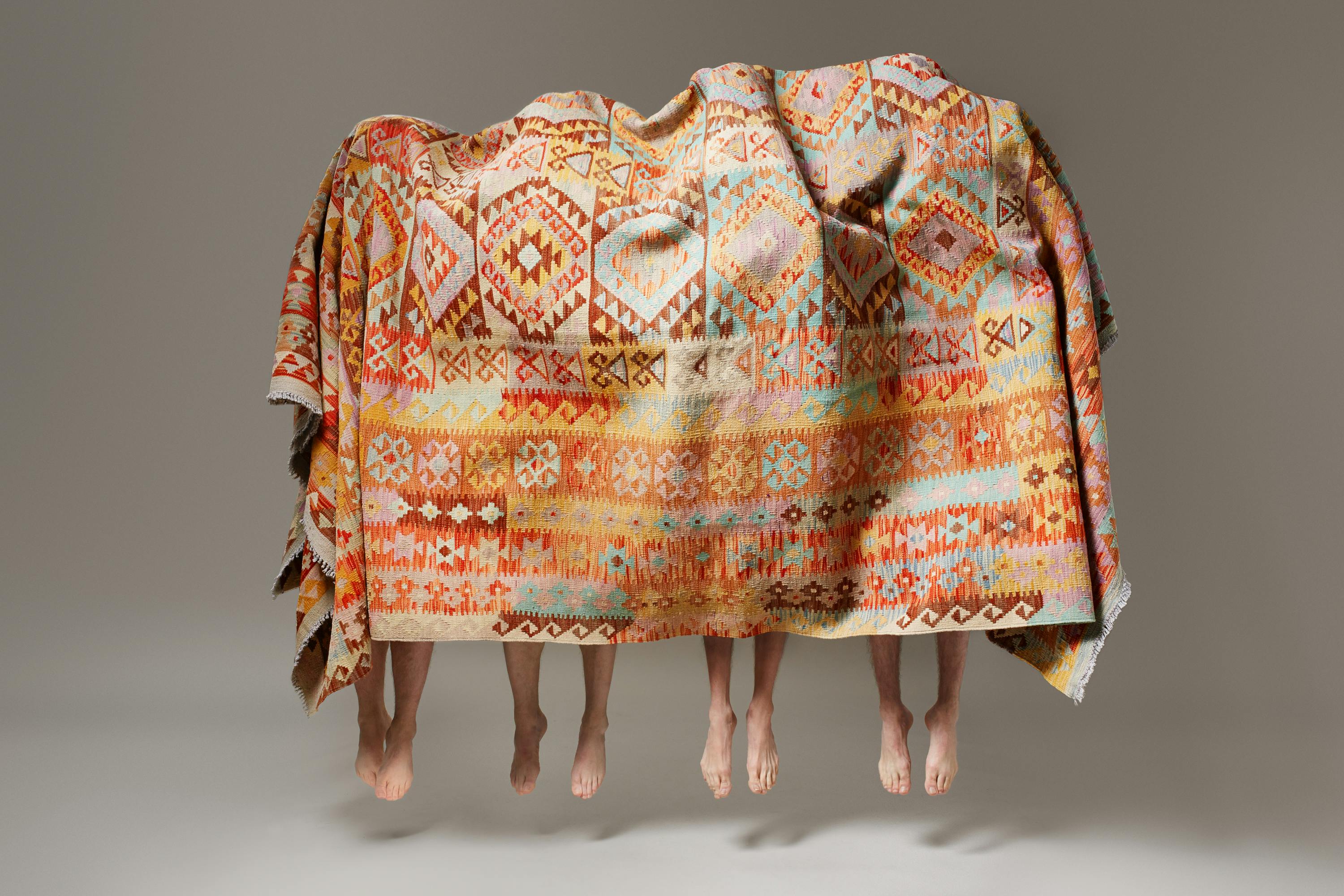 Four men in black trousers under a large carpet all jump into the air at the same time.