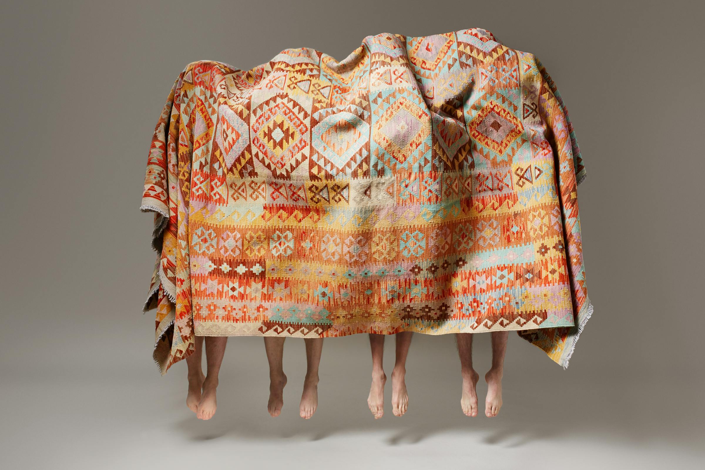 Four men in black trousers under a large carpet all jump into the air at the same time.