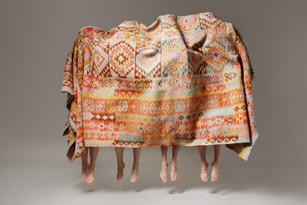 Four men in black trousers under a large carpet all jump into the air at the same time.