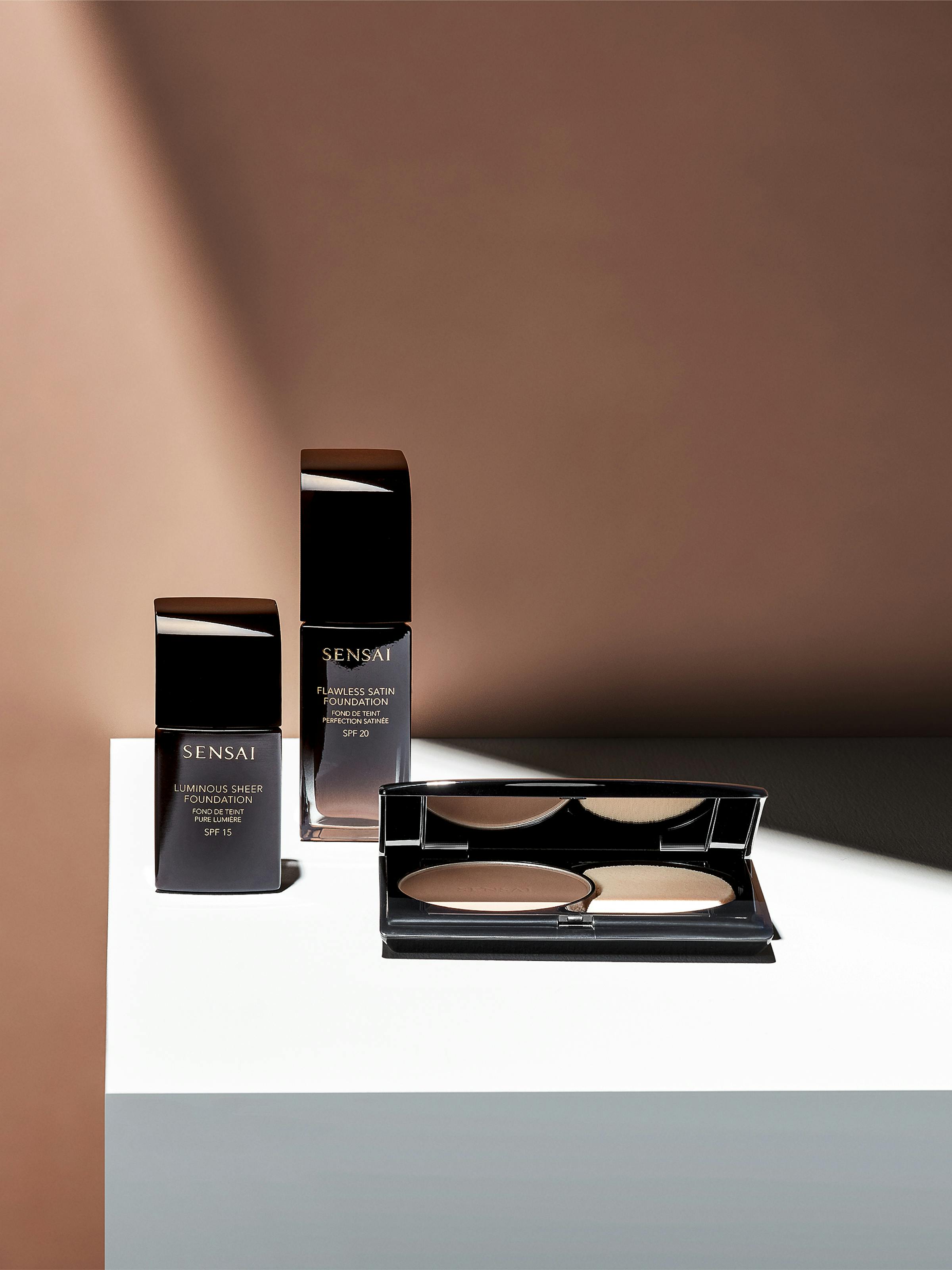 Three dark brown cosmetic products on a grey pedestal in front of a beige wall with strong light incidence.