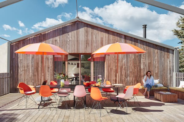 Large wooden terrace with a long black table and Eames shell chairs in orange and pink. A woman is talking on the phone in the background, sitting on a bench.