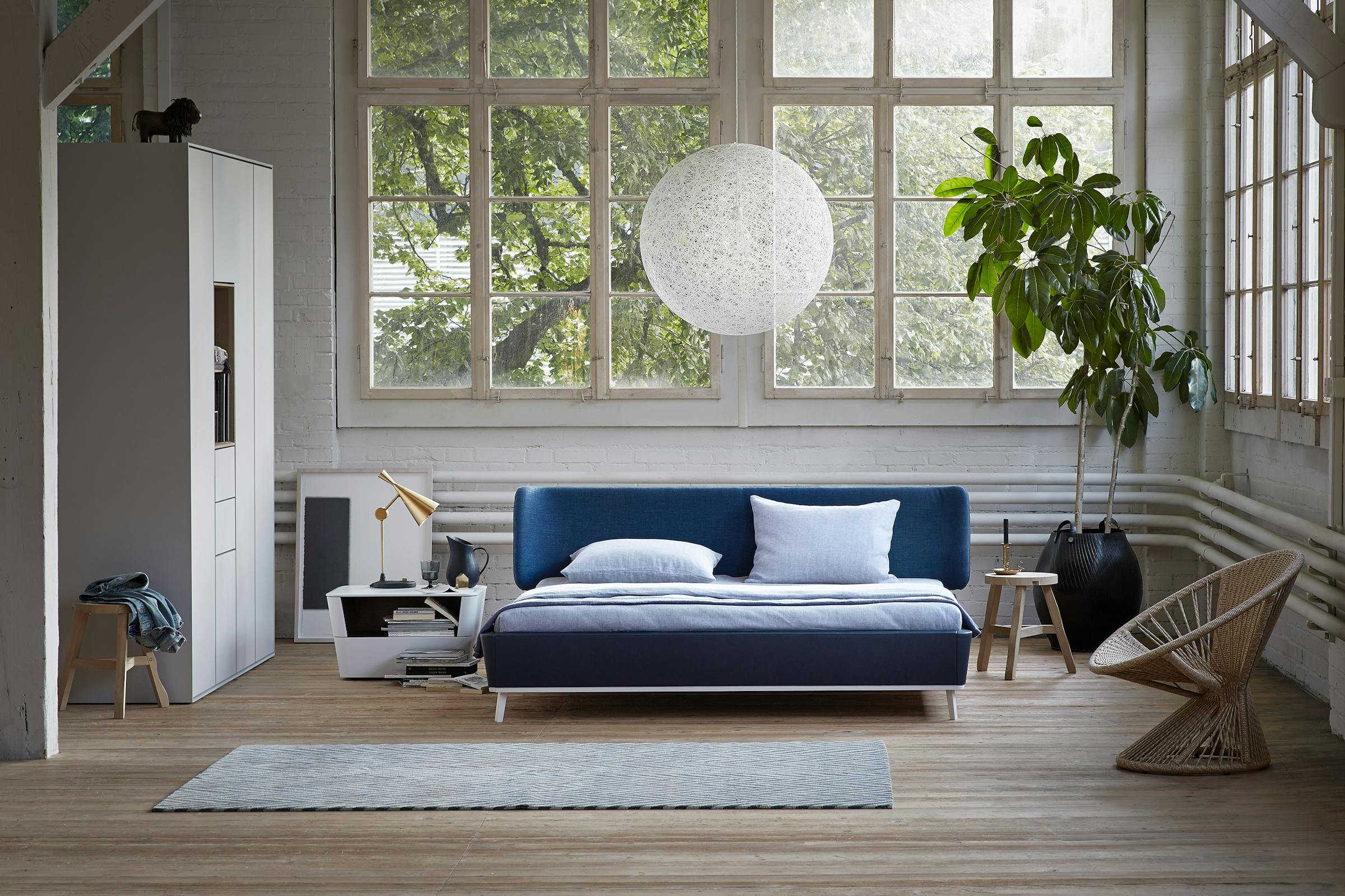 Large dark blue bed in front of a window front in a factory loft on a wooden plank floor