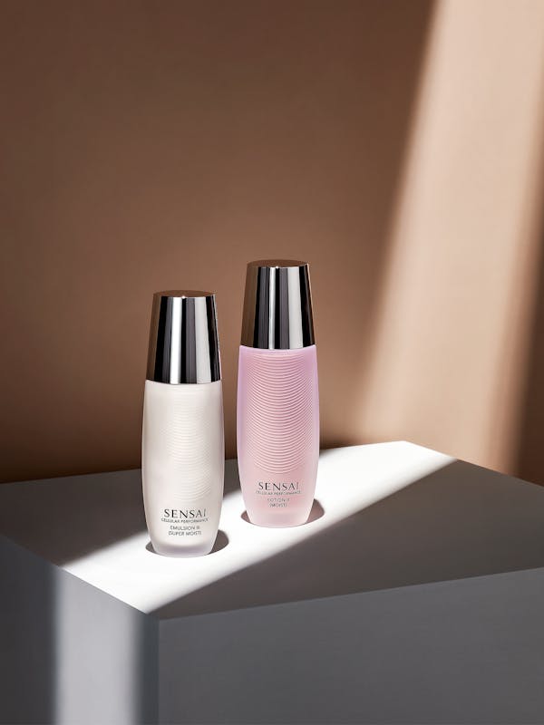 Two glass bottles of beauty creams on a grey pedestal in front of a beige wall with strong light falling on them