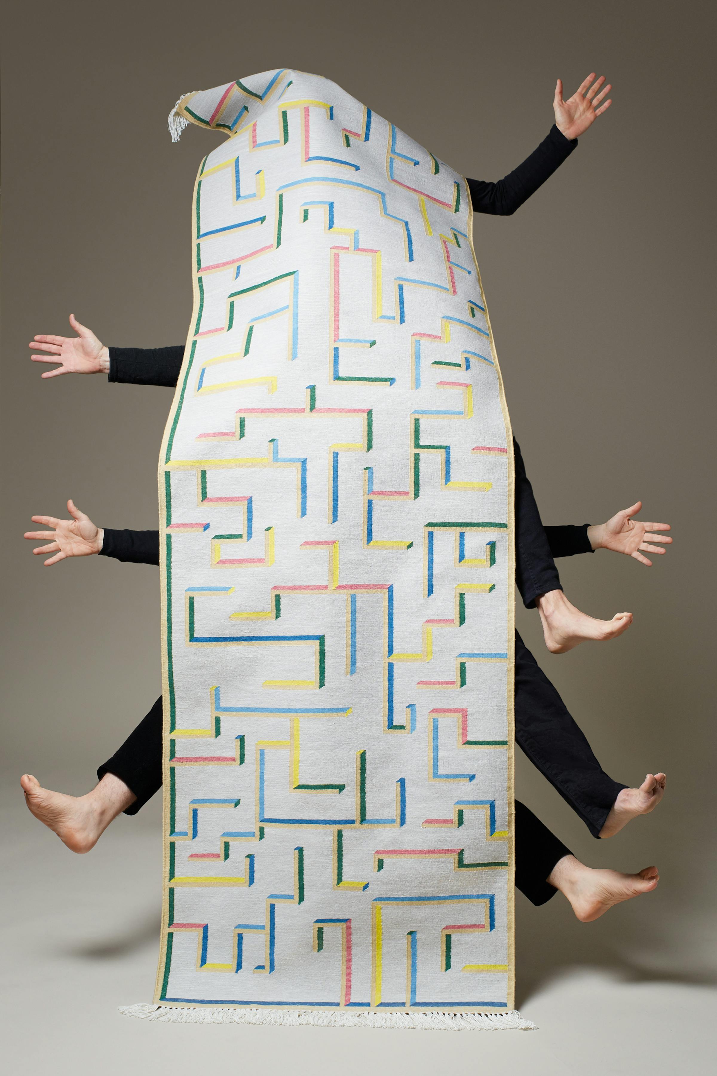 Three men wearing black trousers and black jumpers show only their hands and feet behind a long carpet runner with a graphic pattern.
