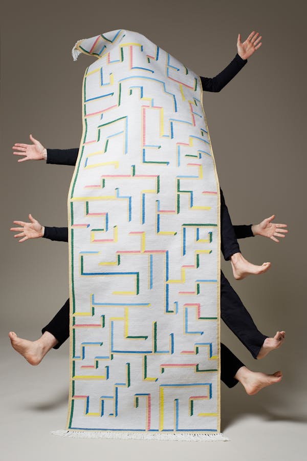 Three men wearing black trousers and black jumpers show only their hands and feet behind a long carpet runner with a graphic pattern.