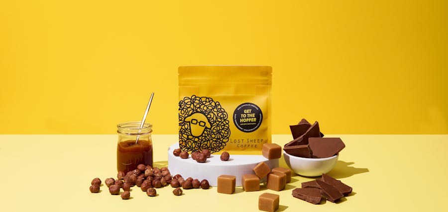 A bag of Lost Sheep Coffee's Get to the Hopper Coffee beans surrounded by caramel, chocolate, fudge and hazelnuts