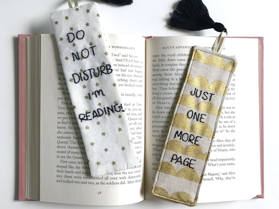 Two fabric bookmarks embroidered in black thread, resting on an open book. One reads 'Do Not Disturb, I'm reading', the second one reads 'Just one more page'