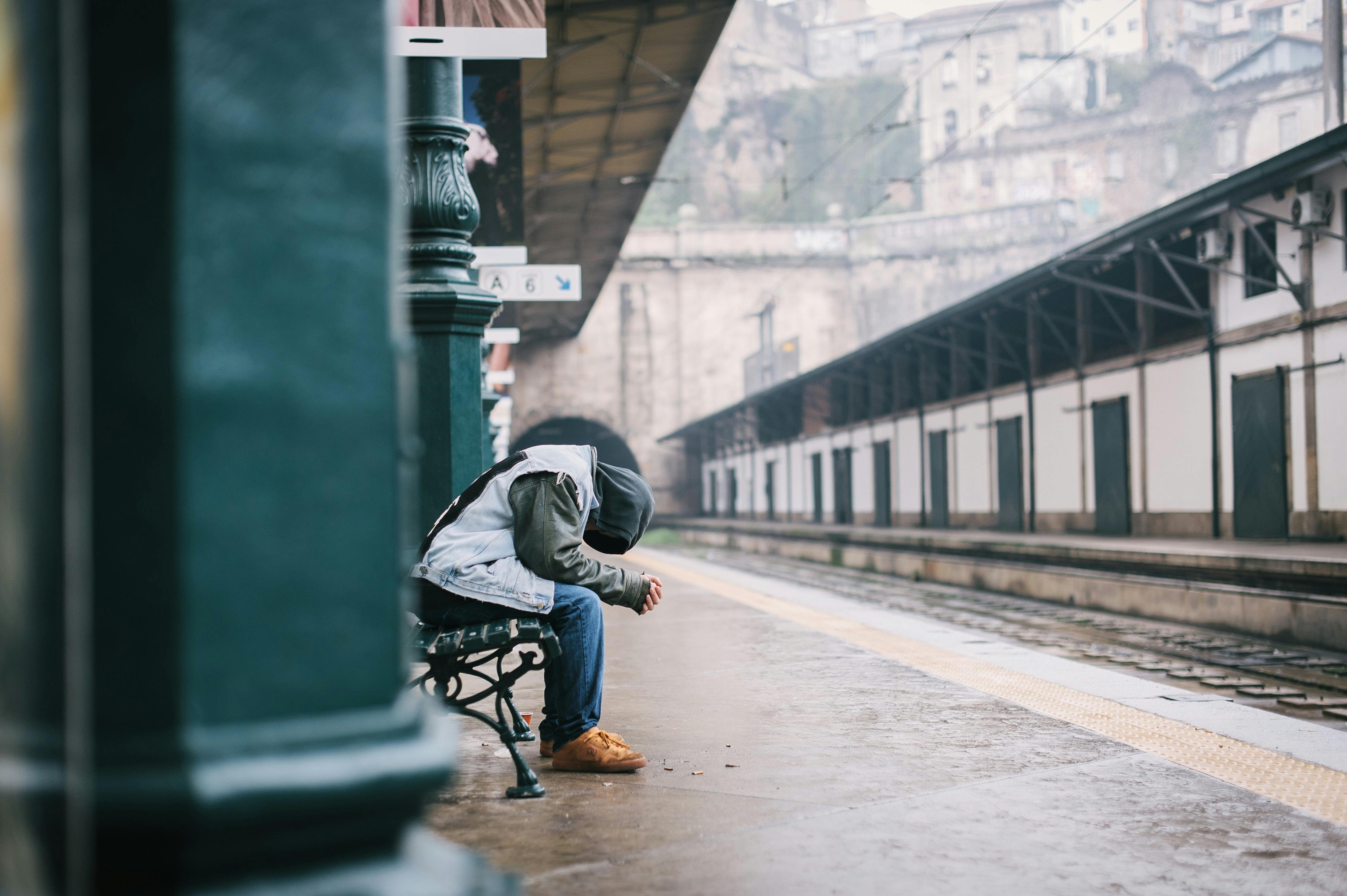 Sad person in hoodie sitting alone on a bench at a dark train station.