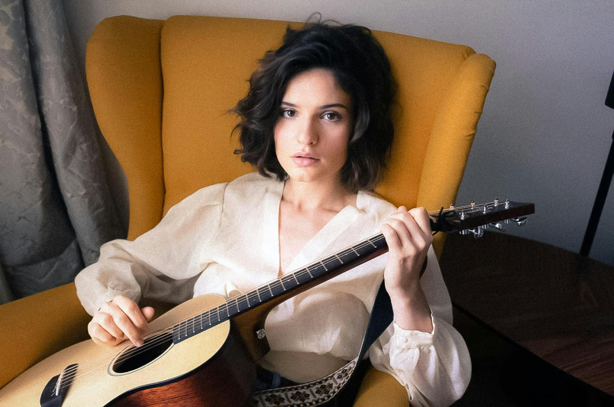 Violetta Zironi sitting in a yellow chair with her acoustic guitar, posing for a photoshoot with jet black hair.