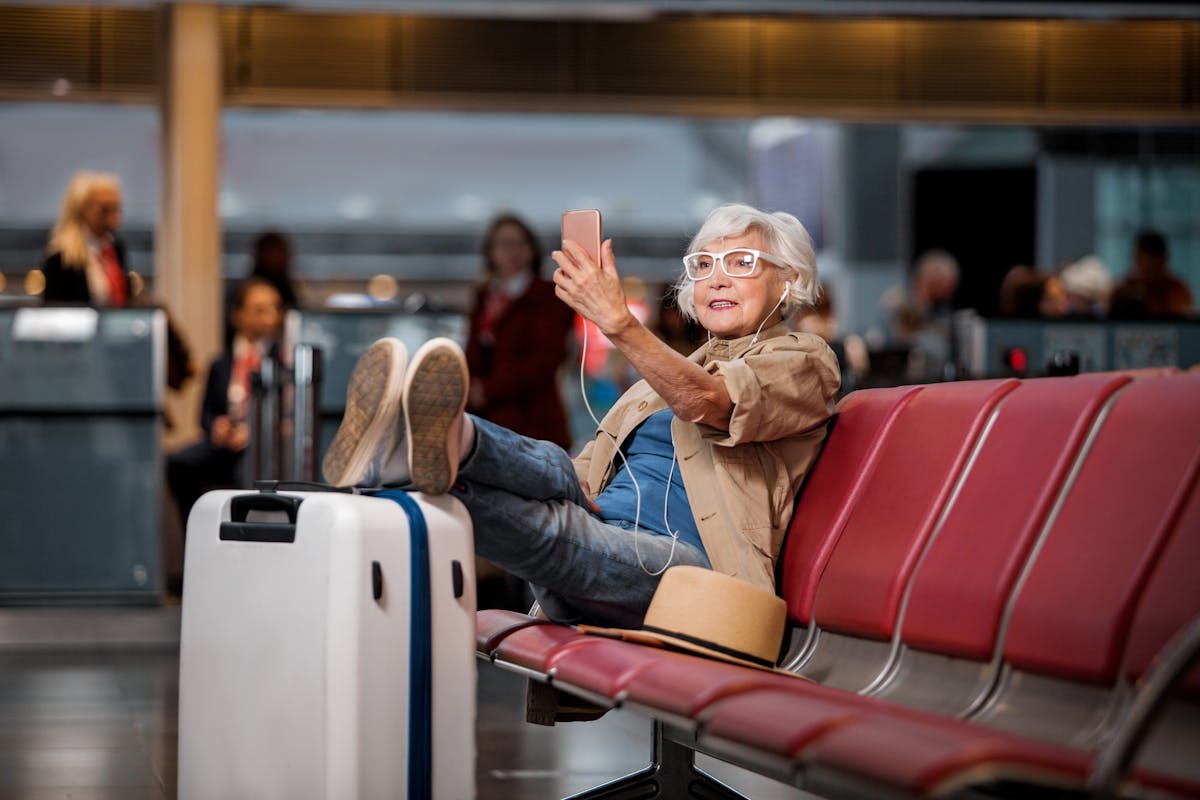 Full length portrait of old lady is sitting on bench at airport lounge. She is having video call using smartphone and earphones while putting legs on suitcase and looking at screen of gadget