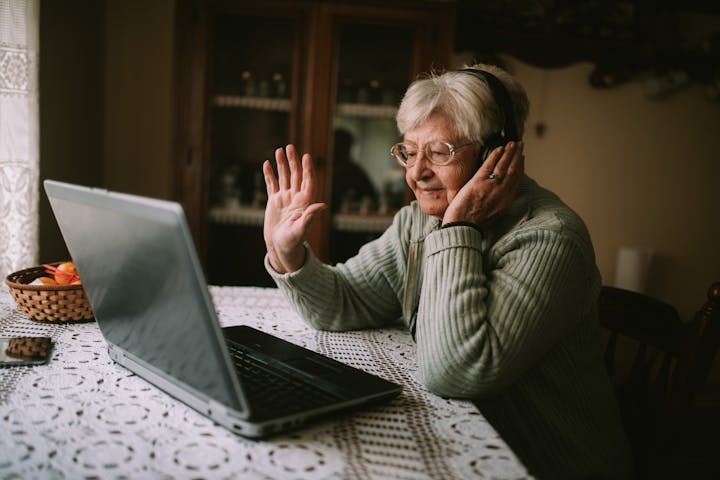 Smiling senior caucasian woman with headphones on her head sitting at a table in front of a laptop and greeting family during quarantine COVID - 19 coronavirus