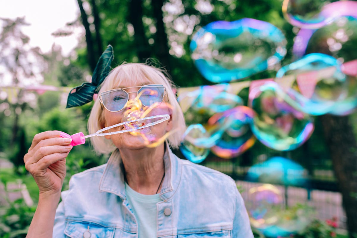 senior (old) stylish woman with gray hair and in blue glasses and jeans jacket blowing bubbles outdoors. Holidays, party, anti age, fun concept