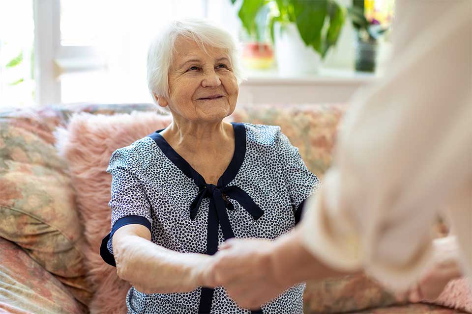 An older woman smiles warmly while holding hands with another person in a comfortable home setting.