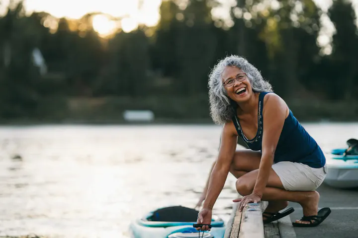 A smiling older woman crouches on a dock beside a calm lake, appearing to prepare a kayak for a peaceful outing at sunset.