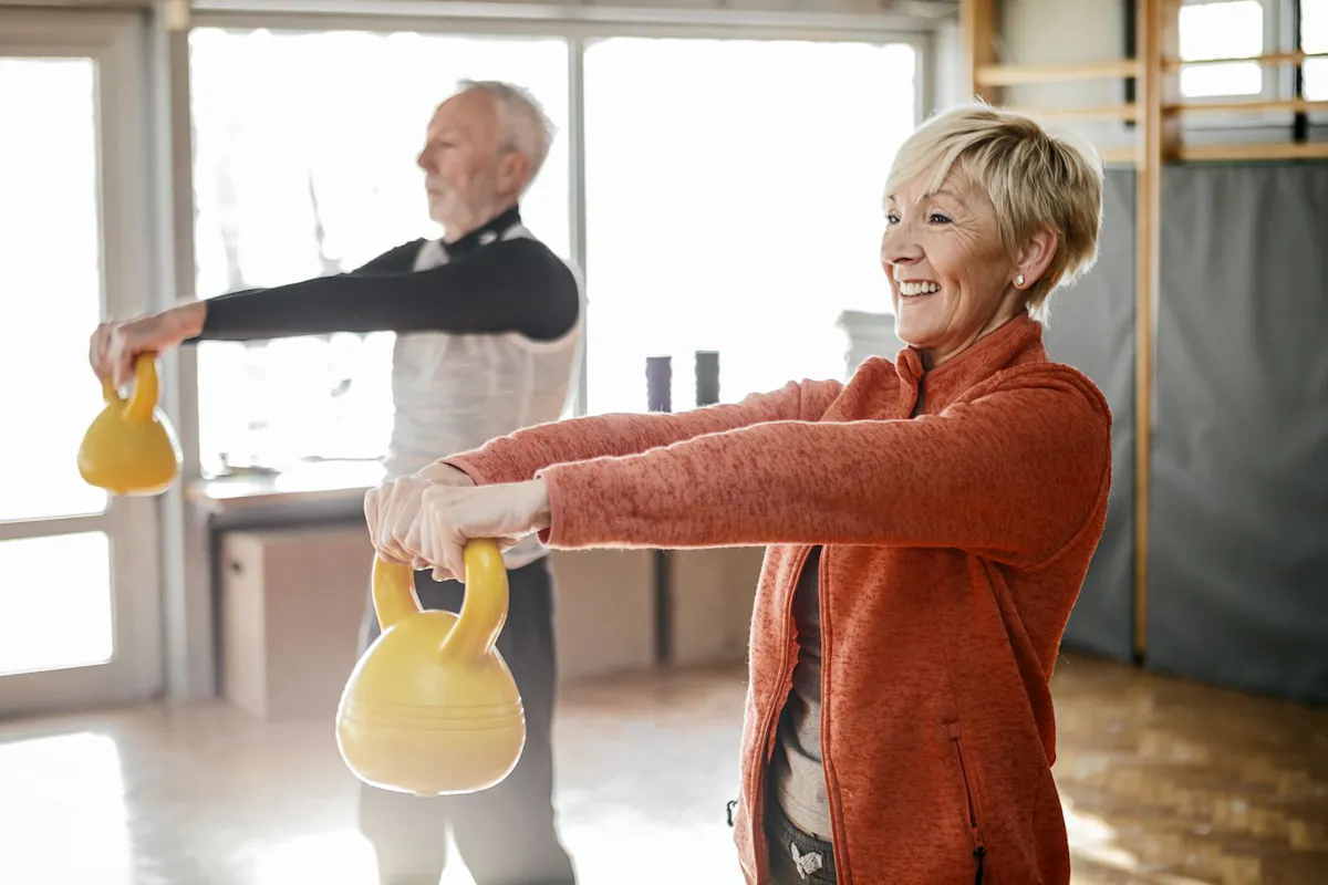 Two older adults practice strength training with yellow kettlebells in a bright fitness studio, promoting healthy aging, muscle strength, and an active lifestyle.