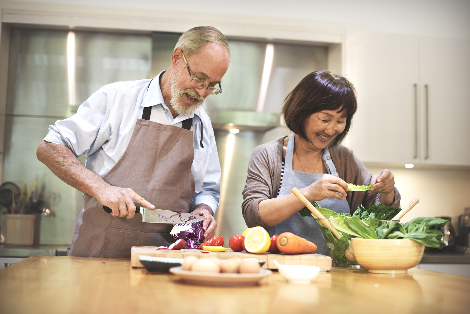 An older couple happily prepares a fresh, colorful meal together in a bright kitchen, surrounded by healthy vegetables and ingredients.