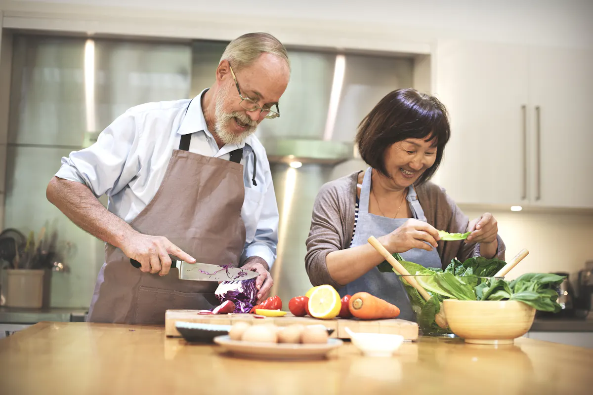 An older couple happily prepares a fresh, colorful meal together in a bright kitchen, surrounded by healthy vegetables and ingredients.